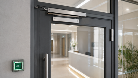 Commercial glazed office door with a monitored magnetic lock on the frame and a green emergency door release on the wall