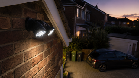 Black twin-head LED security light on the brick gable end of a British semi-detached house, aimed down across a block-paved driveway at dusk
