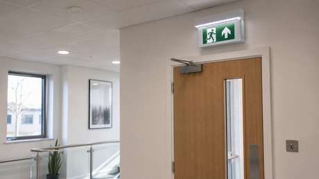White LED emergency exit sign with a running-man legend above a fire door in a UK office corridor, key-switch plate on the wall below