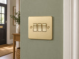 Knightsbridge FP4000BB brushed brass light switch mounted on the wall in a UK living-room entrance wall with muted sage paint.