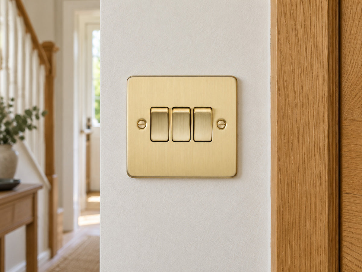 Knightsbridge FP4000BB brushed brass light switch mounted on the wall in a UK hallway wall beside an oak door frame.